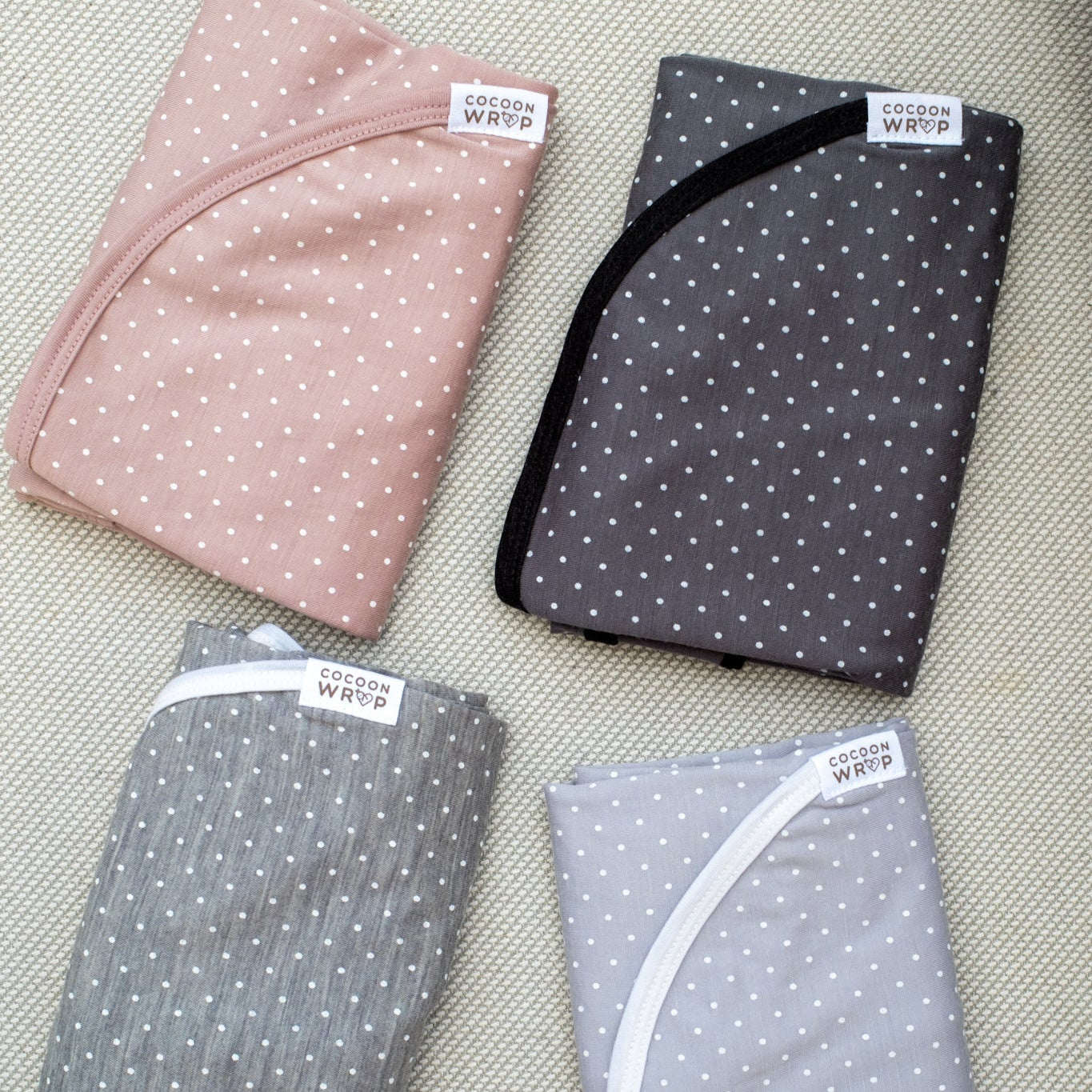 Four folded fabric swaddles in pink, gray, and blue with white polka dots on a textured surface.